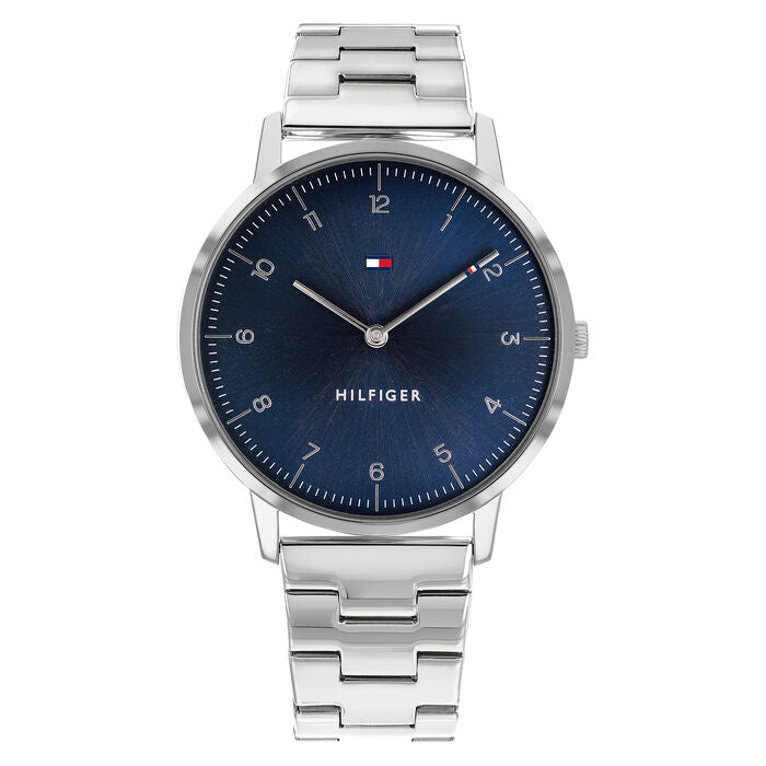A minimalist Tommy Hilfiger men's watch with a 41mm round stainless steel case and a polished silver metal bracelet. The watch features a deep blue sunray dial with small silver Arabic numerals, slim hour markers, and sleek silver hands. The Tommy Hilfiger flag logo is positioned at the 12 o'clock mark, and the word "HILFIGER" is printed above the 6 o'clock position. The design is clean, elegant, and modern.