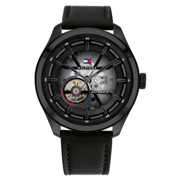 A Tommy Hilfiger men's automatic watch with a 45mm round black stainless steel case and a black leather strap. The watch features a semi-transparent black dial that reveals part of the inner mechanical movement. It includes a visible open-heart complication at the 9 o'clock position, silver-tone hands, and a detailed outer minute track. The Tommy Hilfiger flag logo is placed near the 12 o’clock mark, with the word “AUTOMATIC” printed on the right side of the dial. The design is bold, modern, and mechanical 