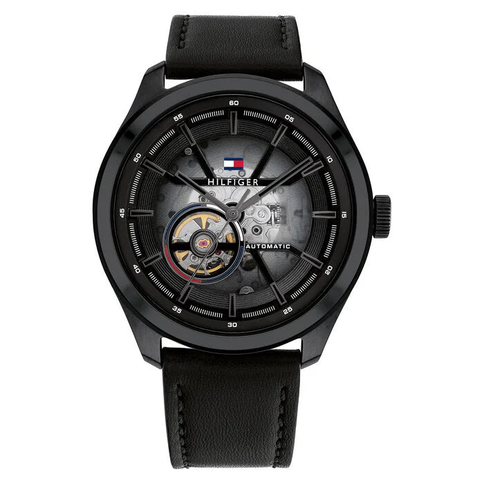 A Tommy Hilfiger men's automatic watch with a 45mm round black stainless steel case and a black leather strap. The watch features a semi-transparent black dial that reveals part of the inner mechanical movement. It includes a visible open-heart complication at the 9 o'clock position, silver-tone hands, and a detailed outer minute track. The Tommy Hilfiger flag logo is placed near the 12 o’clock mark, with the word “AUTOMATIC” printed on the right side of the dial. The design is bold, modern, and mechanical 