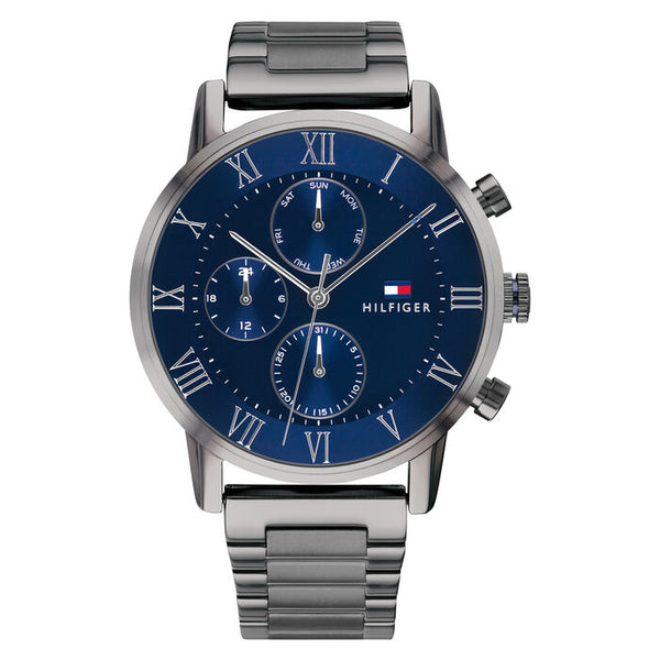 A Tommy Hilfiger men's watch with a 44mm round stainless steel case and a polished gunmetal bracelet. The watch features a deep blue dial with silver-tone Roman numeral hour markers and three subdials showing day of the week, date, and 24-hour time. It includes silver-tone hands and a slender second hand, with the Tommy Hilfiger flag logo and "Hilfiger" branding on the right side of the dial. The design is sleek, modern, and sophisticated.