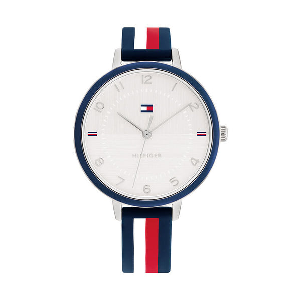 A Tommy Hilfiger women's watch with a 38mm stainless steel case and a navy blue bezel. The white dial features subtle textured detailing with silver-tone hands, Arabic numerals, and small Tommy Hilfiger flag accents at the 3, 6, 9, and 12 o'clock positions. The strap is navy blue silicone with bold vertical red and white stripes running down the center, reflecting the brand's signature color scheme. The design is sporty, youthful, and modern.