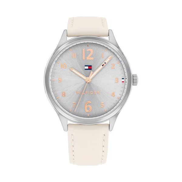 A Tommy Hilfiger women's watch with a 38mm silver-tone stainless steel case and a soft beige leather strap with matching stitching. The dial features a subtle sunburst pattern in silver with rose gold-tone Arabic numerals, hands, and markers. The Tommy Hilfiger flag logo is placed just below the 12 o’clock position, with the word “HILFIGER” above the 6 o’clock mark. The design is feminine, elegant, and minimalistic.